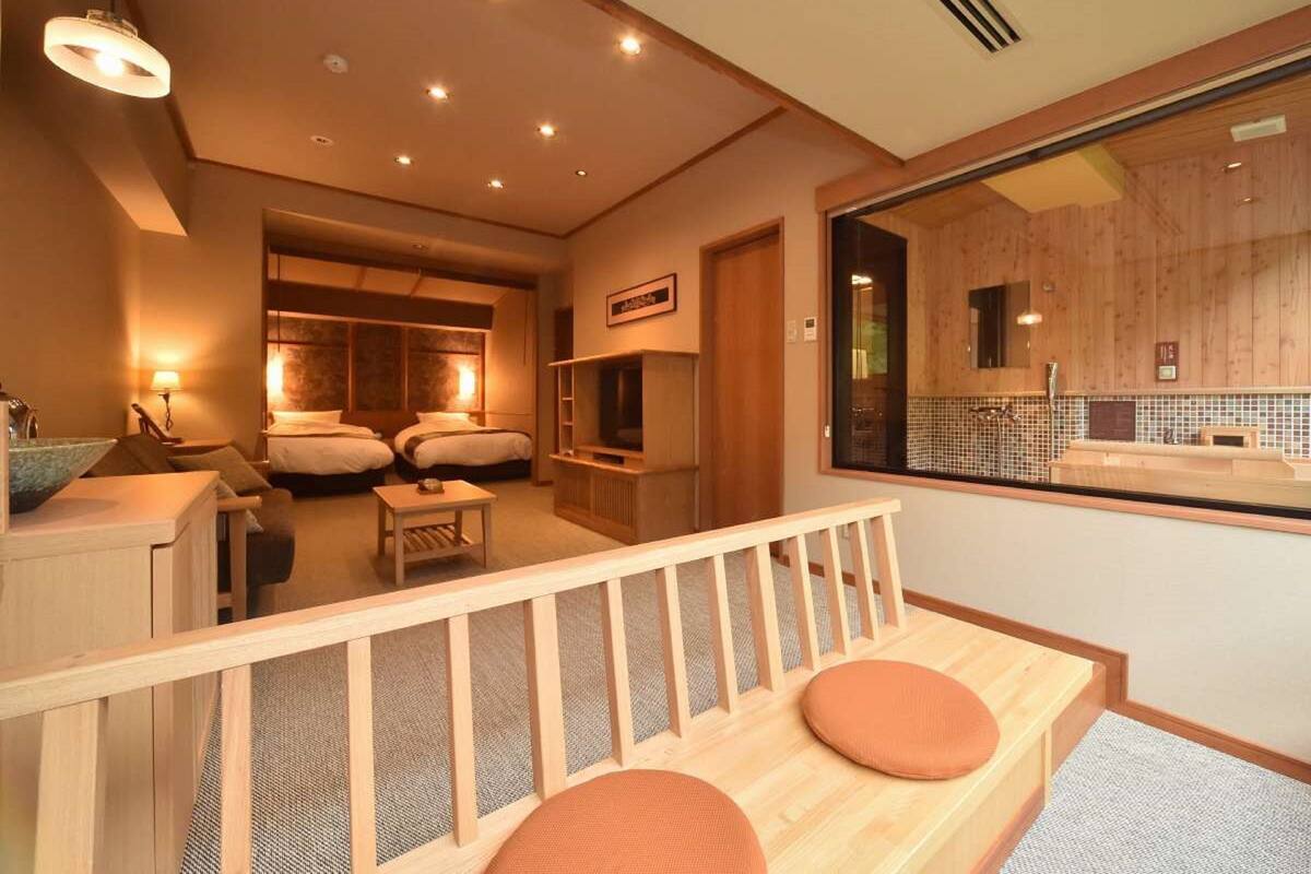 A hotel room with a wooden bench, a TV, and a large window overlooking the bathroom.