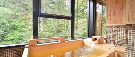 A wooden cabin with a large bathtub filled with water, a wooden bench, and a view of a forest through large windows.