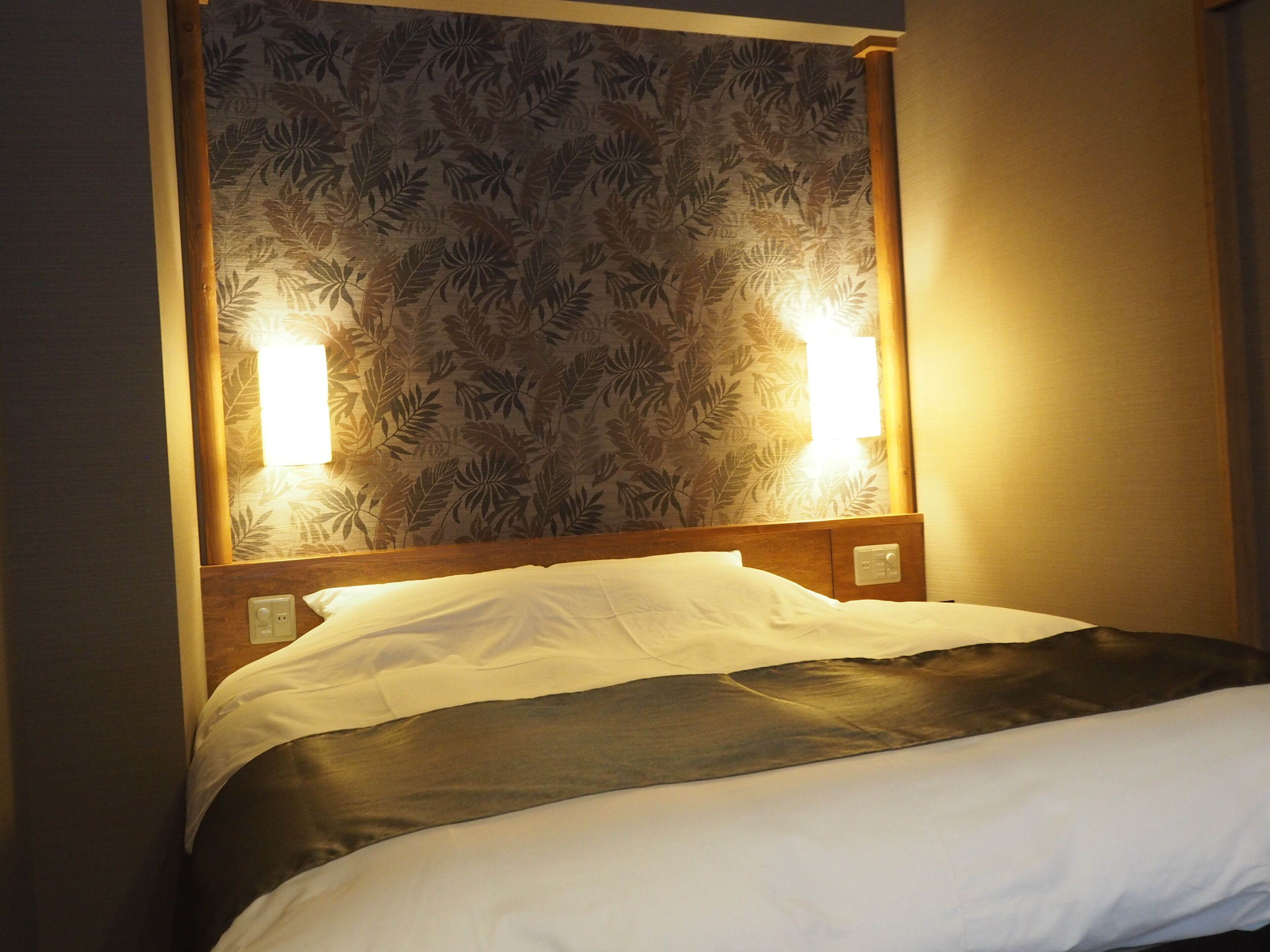 A hotel room with a bed, a headboard with a floral pattern, and two wall-mounted lights.