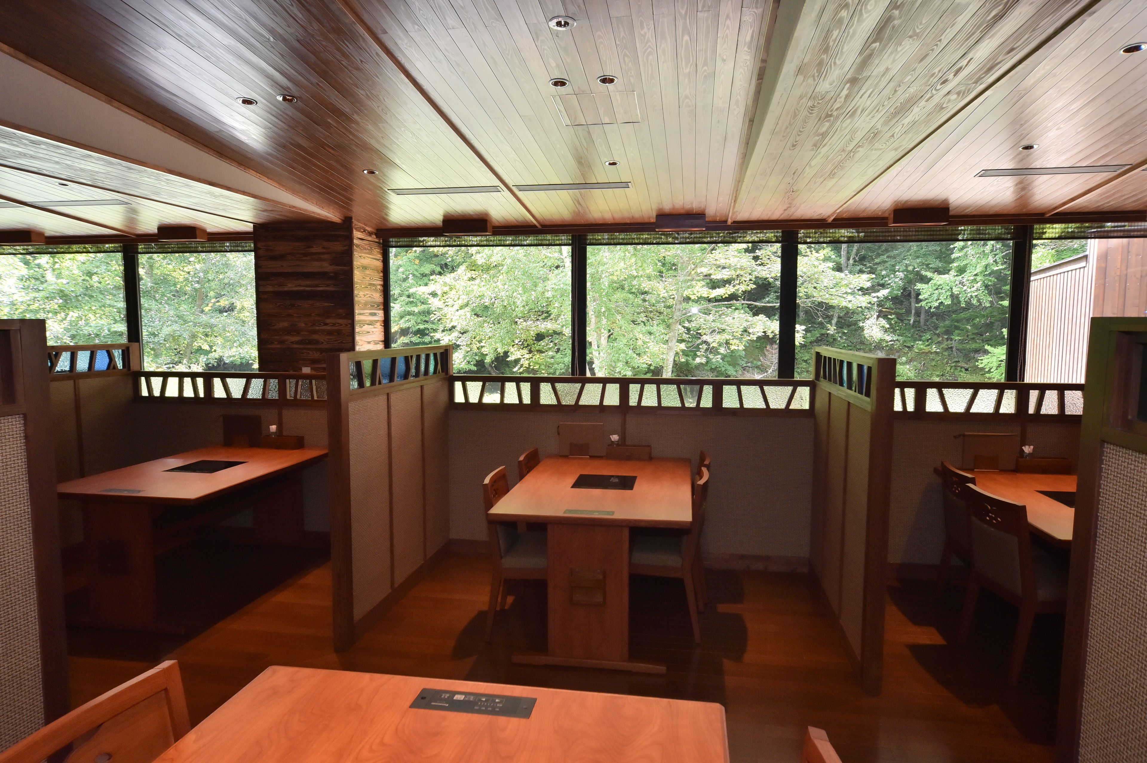 A dining area with wooden tables and chairs, large windows with a view of trees, and a wooden ceiling.
