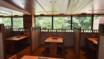 A dining area with wooden tables and chairs, large windows with a view of trees, and a wooden ceiling.