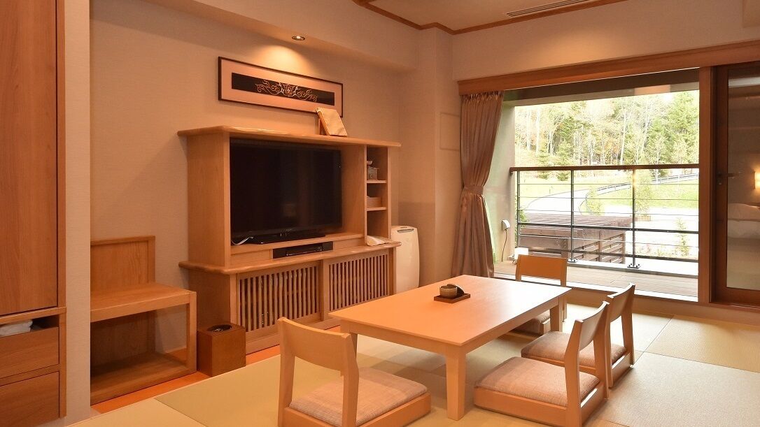 A room with a television, a table with chairs, a window with a view of a road, and a framed picture on the wall.