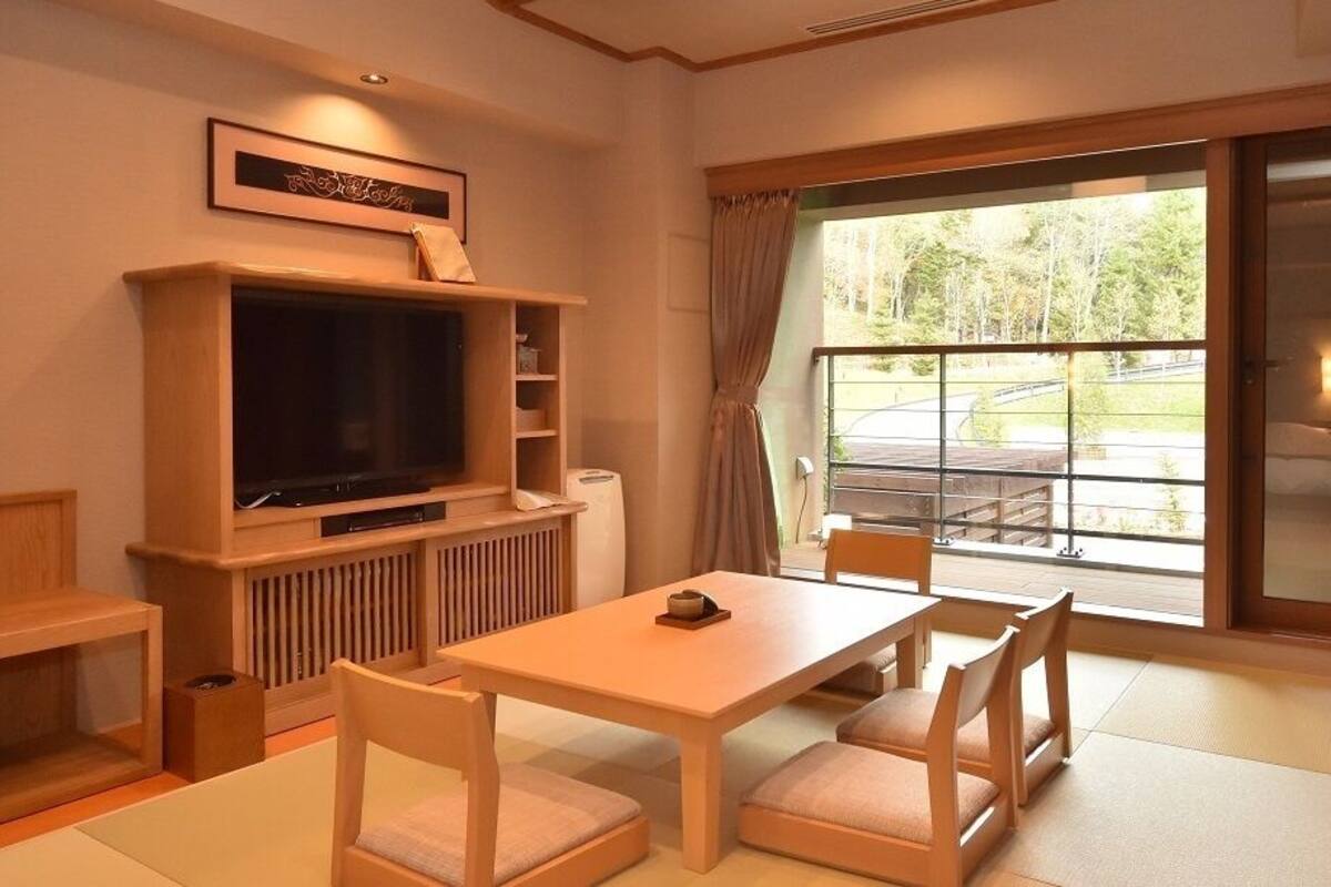 A room with a television, a table with chairs, a window with a view of a road, and a framed picture on the wall.