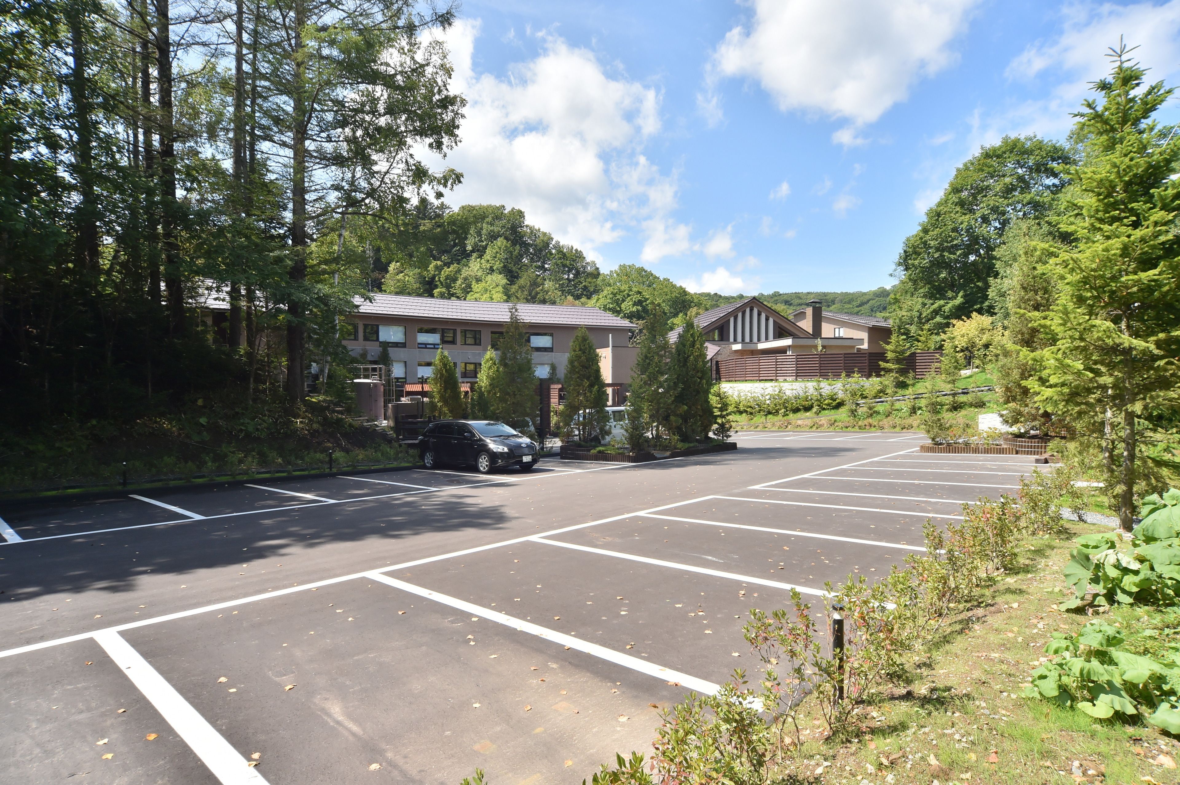 A parking lot with multiple parking spaces, a building with a sloped roof, and surrounding trees.
