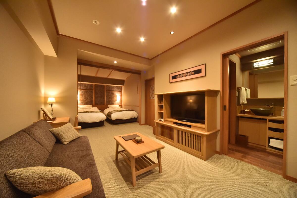 A hotel room with a sofa, a TV, a small table, a bed, and a bathroom visible through an open door.