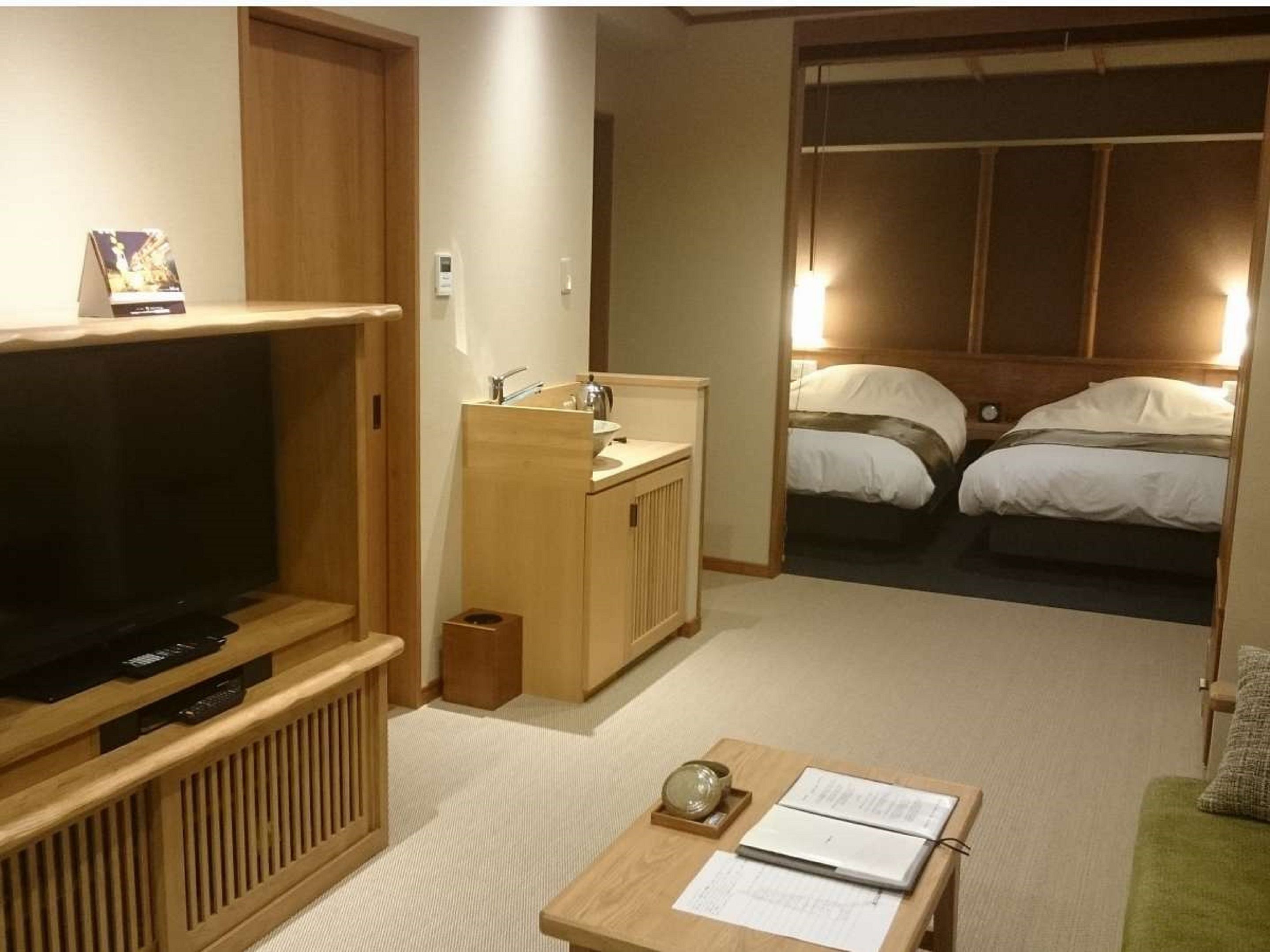 A hotel room with a television, a small kitchenette, a bed, and a seating area with a table and papers.
