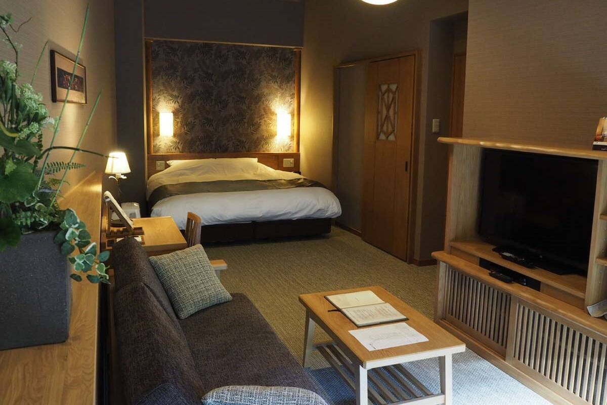 A hotel room with a bed, a TV, a sofa, a small table, and a potted plant.