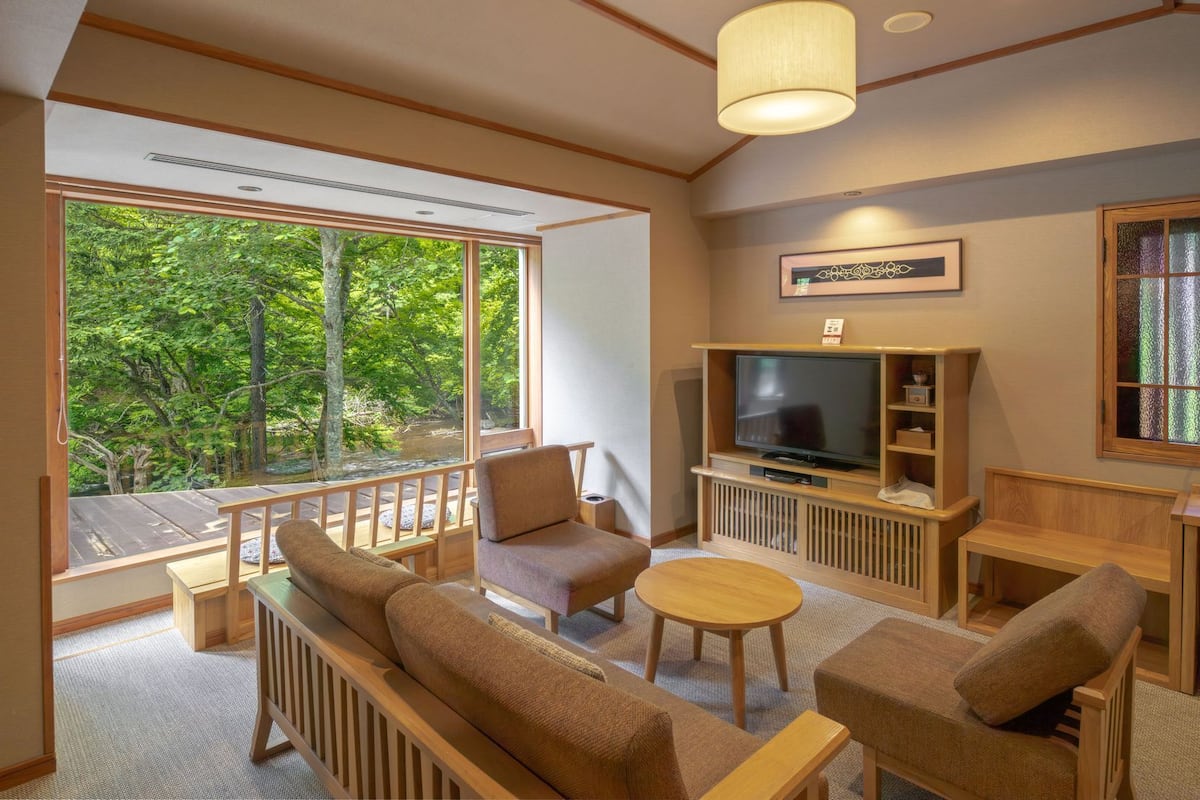 A living room with a TV, sofa, and a view of a forest through large windows.