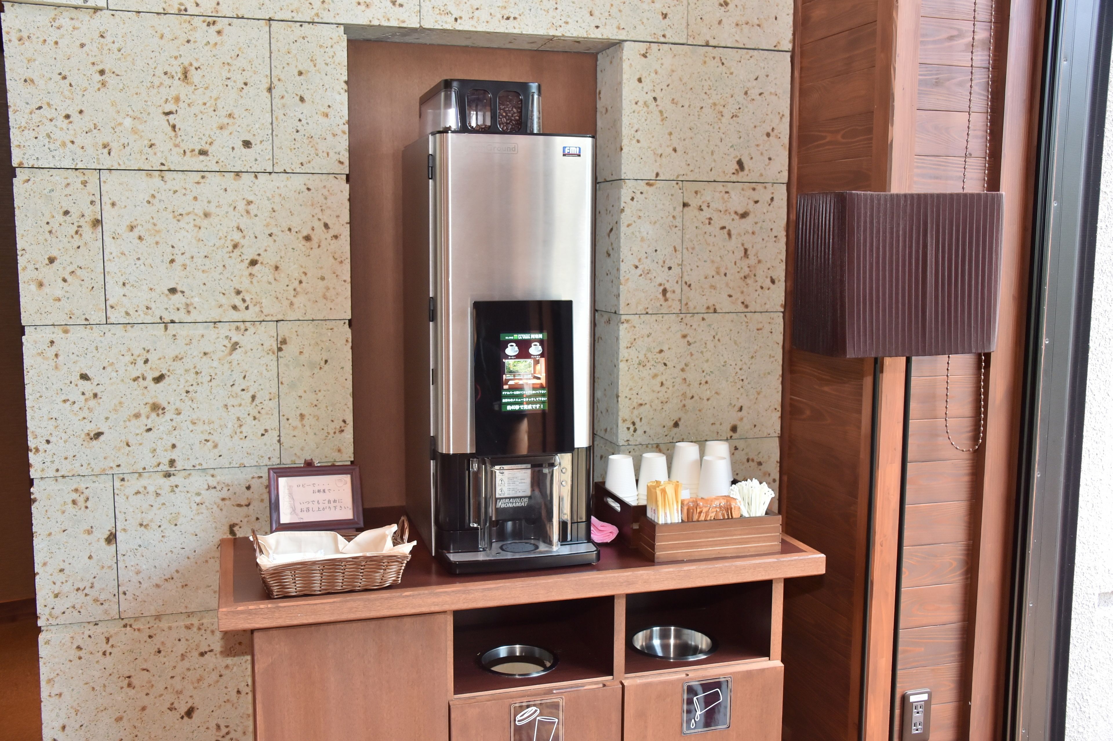 Una macchina del caffè con display digitale, un cesto con tovaglioli e un vassoio con tazze e posate su un bancone in legno.