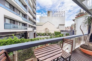 Un balcón con mesa y sillas de madera, una planta en maceta y vistas a un edificio de fachada blanca y un ventanal grande.