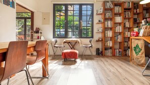 Una habitación con un escritorio de madera, dos sillas, una mesita, una estantería y un ventanal con vistas a árboles.