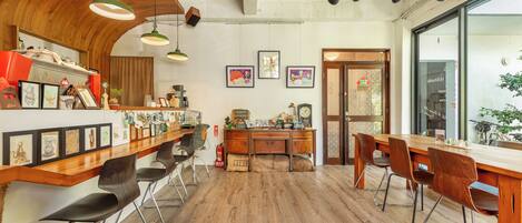 A spacious room with wooden floors, a long bar area, and a dining table with chairs. There are framed pictures on the walls and a large window providing a view outside.