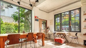 Una habitación amplia con una gran mesa de madera y sillas, una estantería y ventanas grandes que ofrecen vistas a la vegetación exterior.