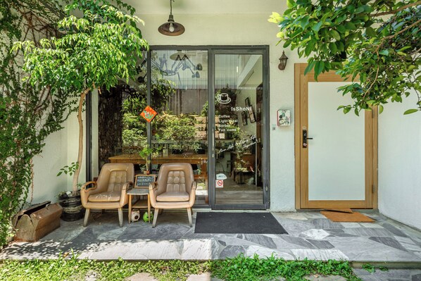 Entrada de un café con puerta de cristal, dos sillas y una mesita en el exterior, y una planta en maceta.