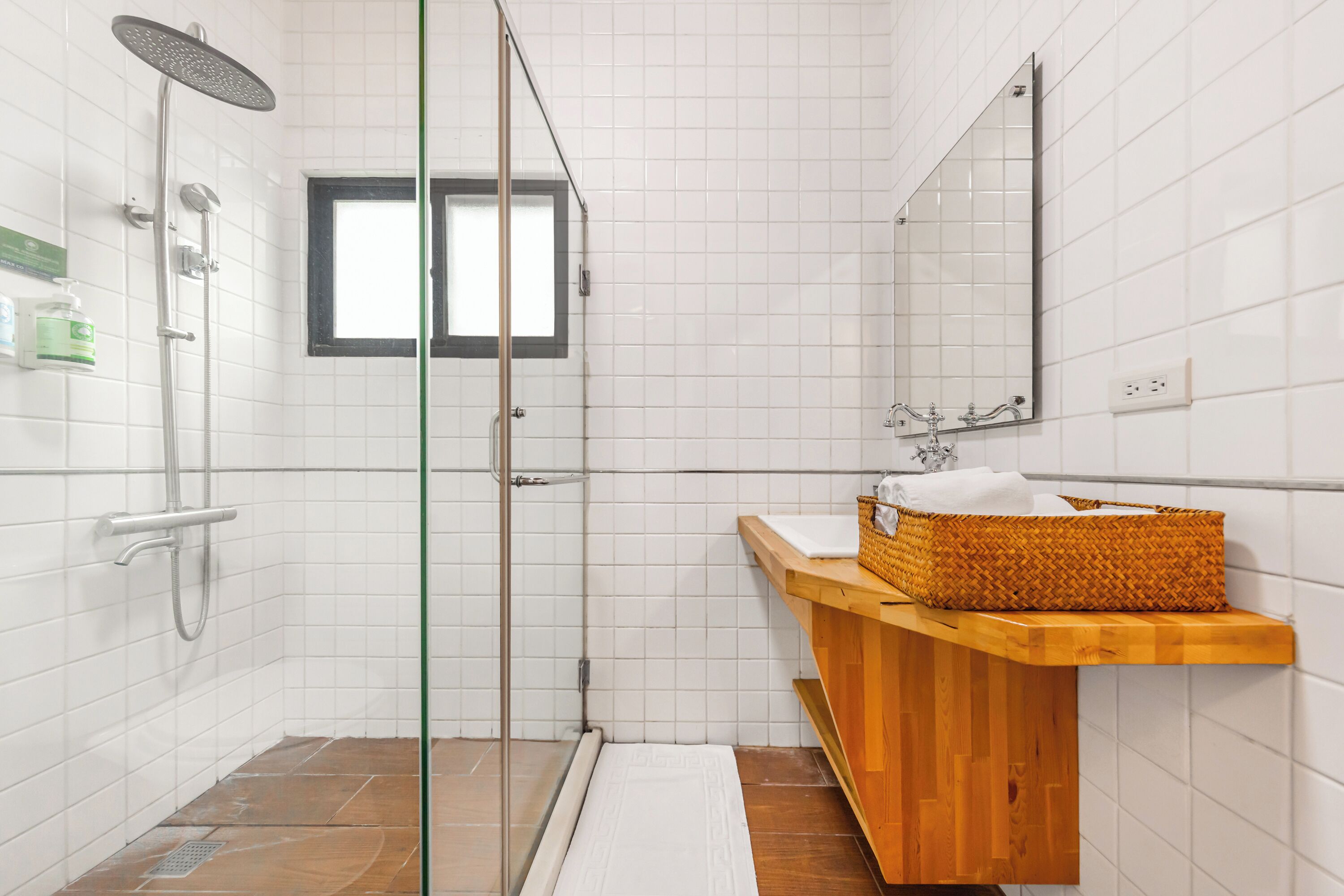 A bathroom with a glass-enclosed shower, a wooden vanity with a basket, and a mirror above the sink.