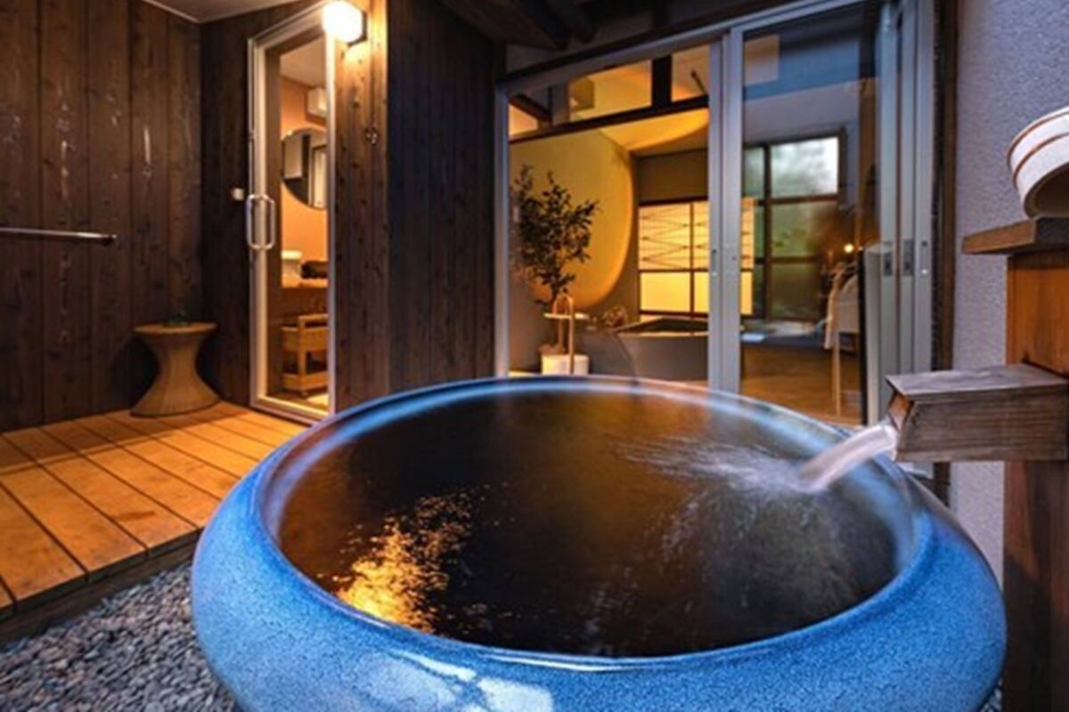 A hot spring bath area with a large stone basin, wooden walls, and a sliding glass door leading to a room with a bathtub and a plant.