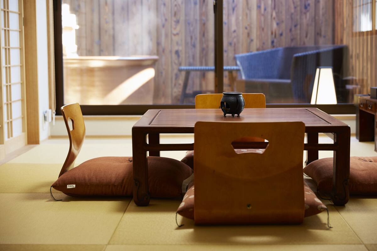 A traditional tatami mat room with a wooden table, cushions, and a view of a tatami mat room through a glass door.