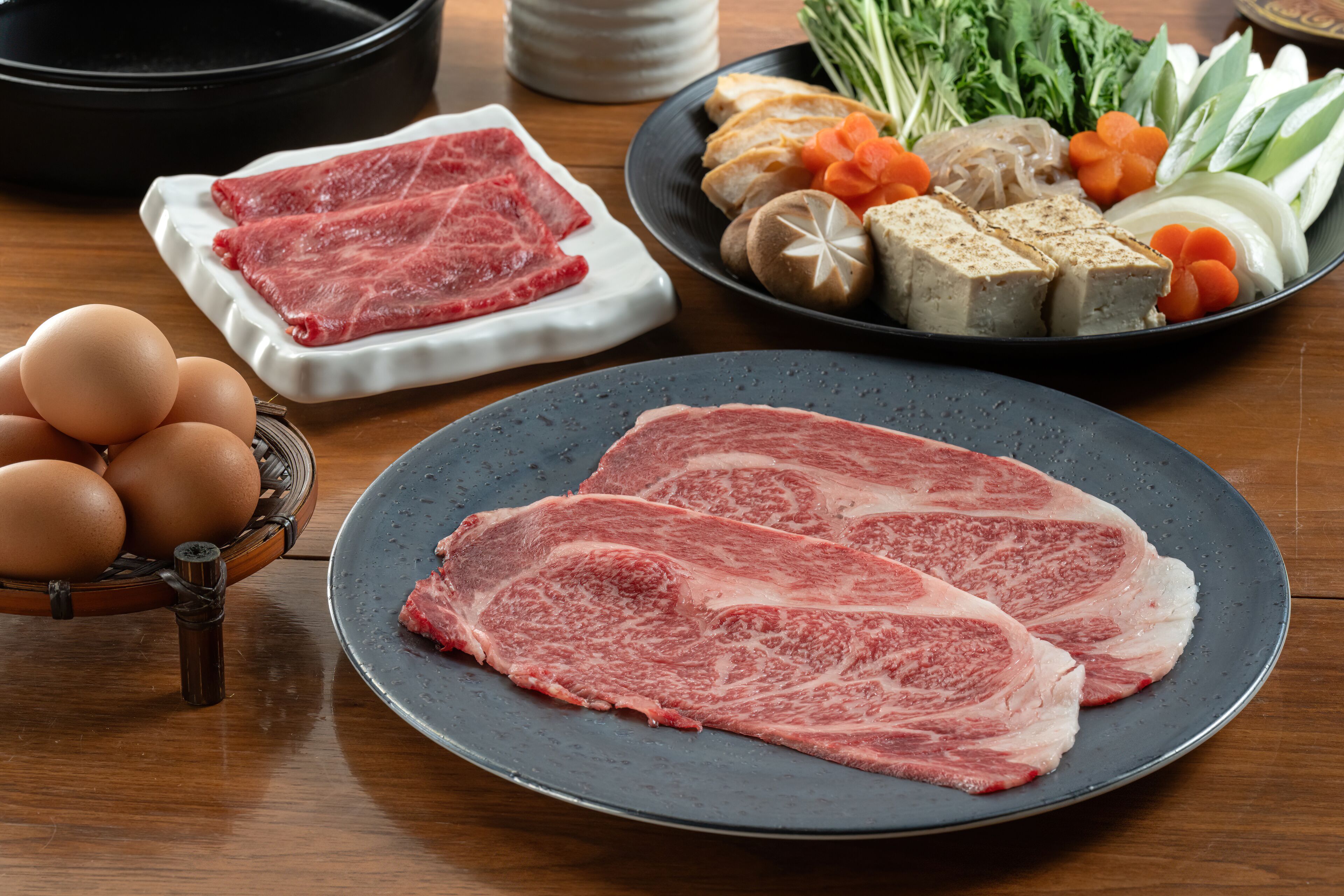 A dining table with various food items including raw meat, eggs, vegetables, and tofu.