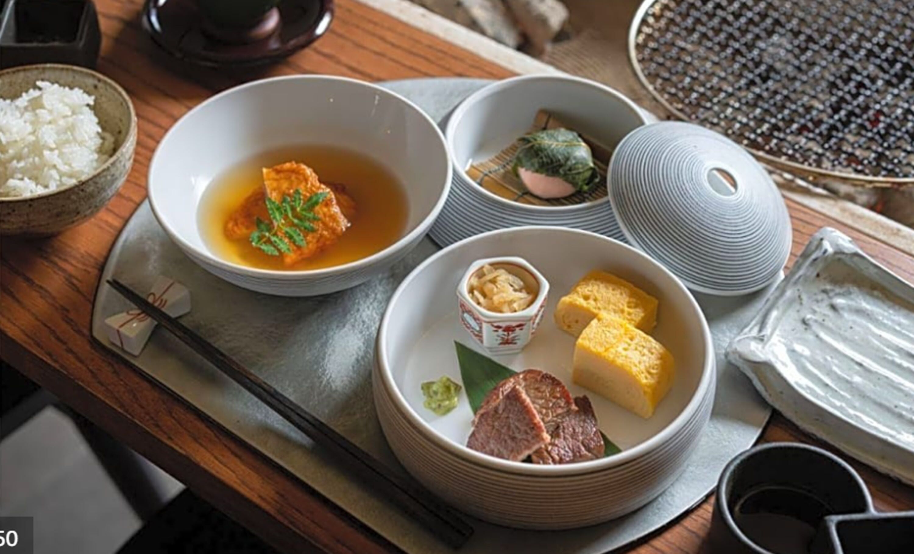 Presentació tradicional de menjar japonès amb diversos plats, com ara arròs, sopa i carn, damunt un taula de fusta.