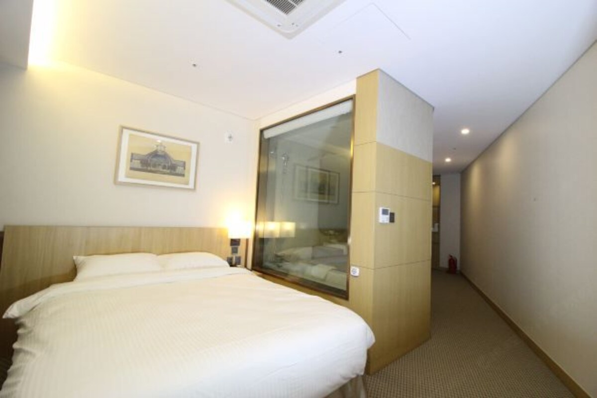 A hotel room with a large bed, a glass partition wall, and a framed picture on the wall.
