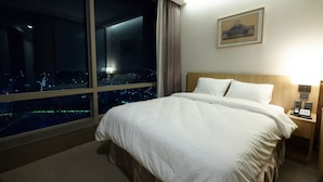 Una habitación de hotel con una cama grande, una mesita de noche con una lámpara, un cuadro enmarcado en la pared y vistas a un paisaje urbano iluminado por la noche a través de un ventanal amplio.