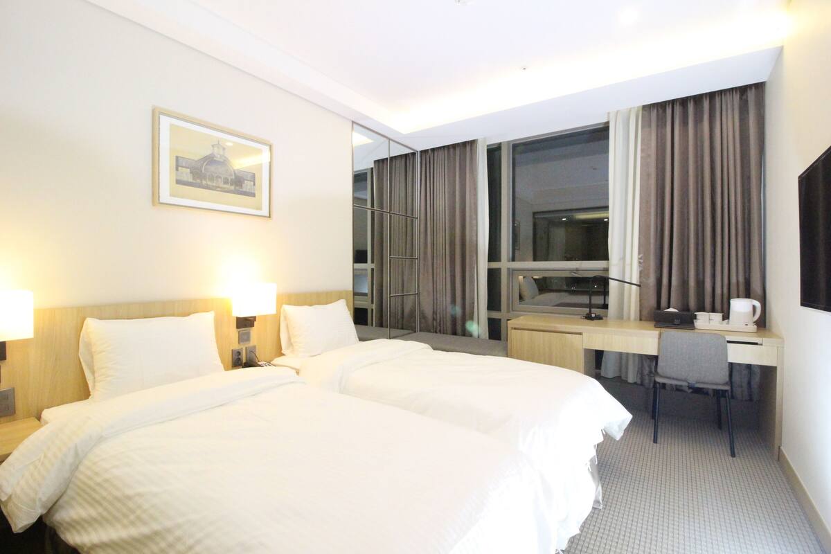 A hotel room with two beds, a desk, a chair, a large window with curtains, and a framed picture on the wall.
