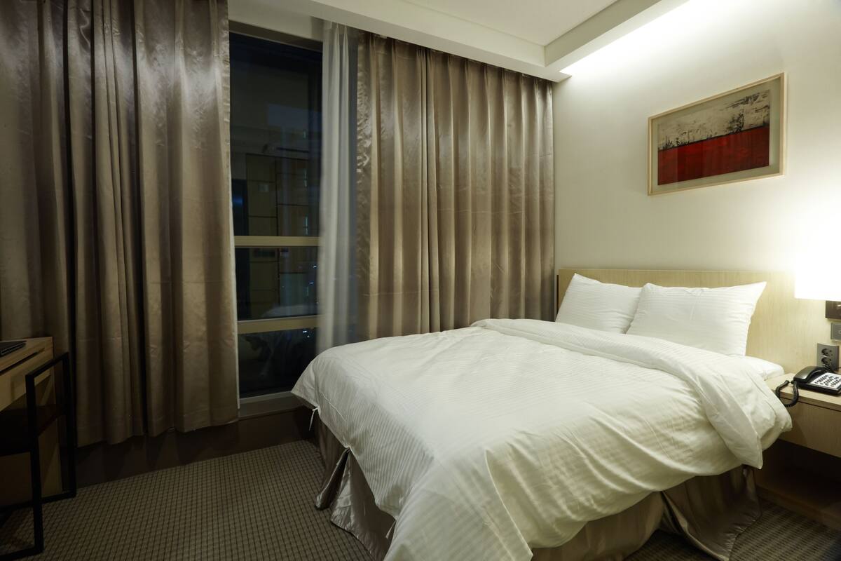 A hotel room with a large bed, a desk, a chair, and a window with curtains.