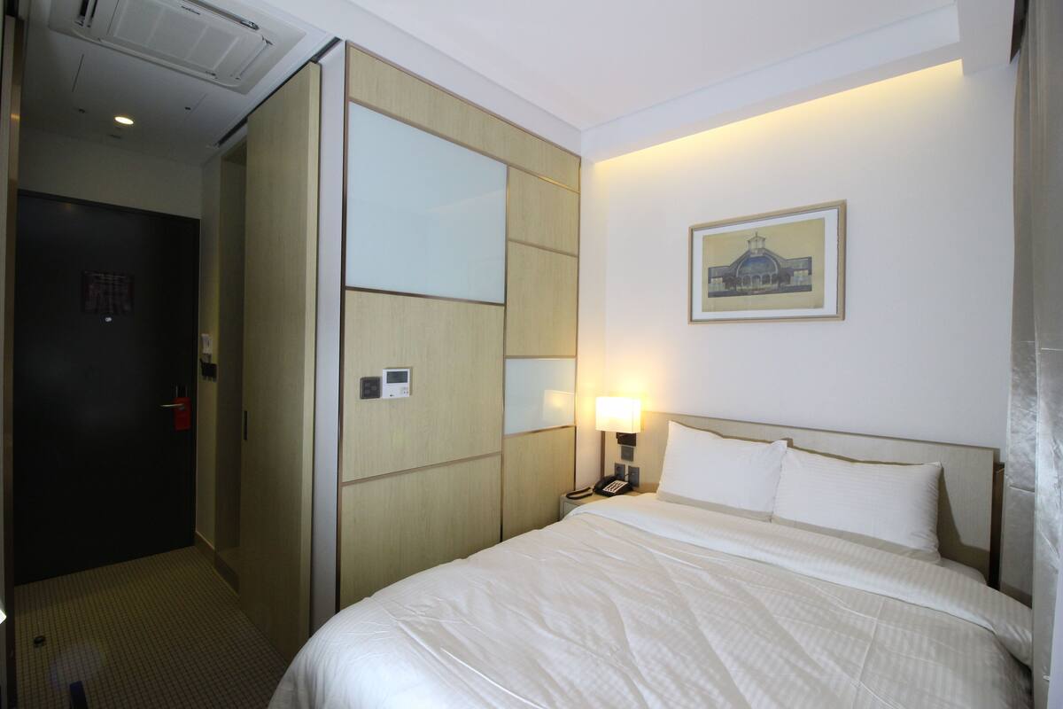 A hotel room with a bed, a nightstand, a lamp, a wall-mounted phone, and a framed picture on the wall.