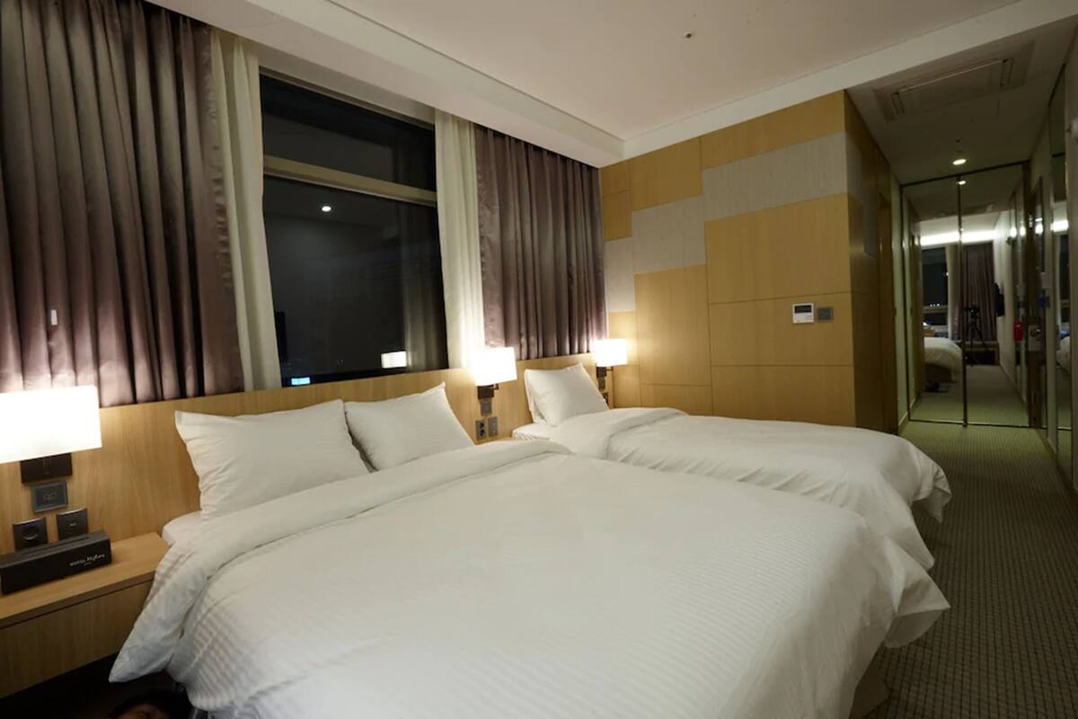 A hotel room with two beds, a nightstand with a lamp, a large window with curtains, and a wall-mounted control panel.