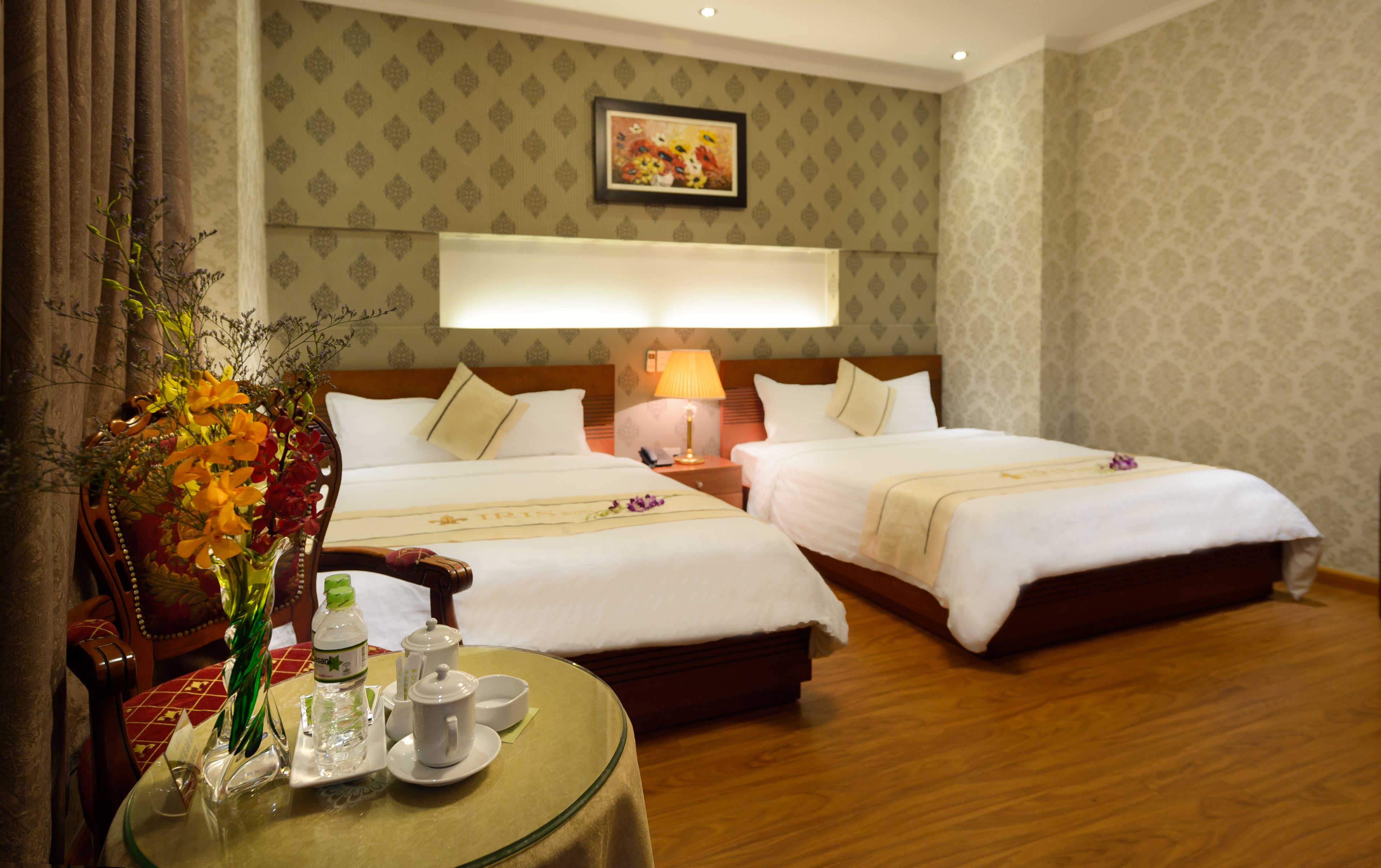 A hotel room with two beds, a bedside table with a lamp, a vase with flowers, and a table with a water bottle and cups.