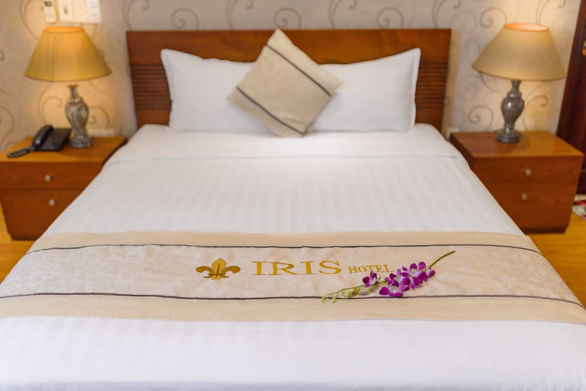 Una cama de hotel con una colcha que muestra el logotipo del HOTEL IRIS y un símbolo de lis.