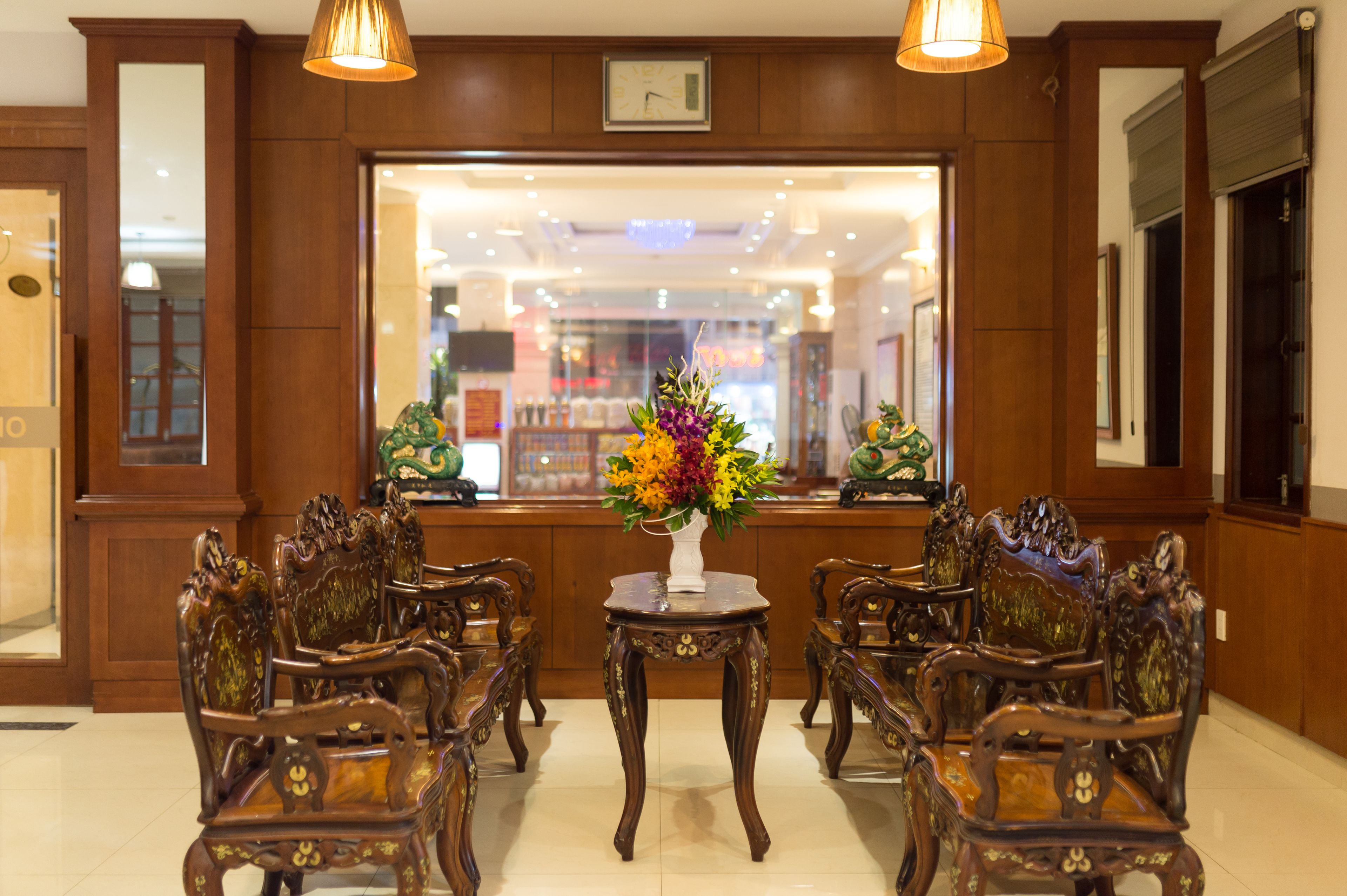 A lobby with ornate wooden furniture, a central table with a vase of flowers, and a large window with a view of the interior.