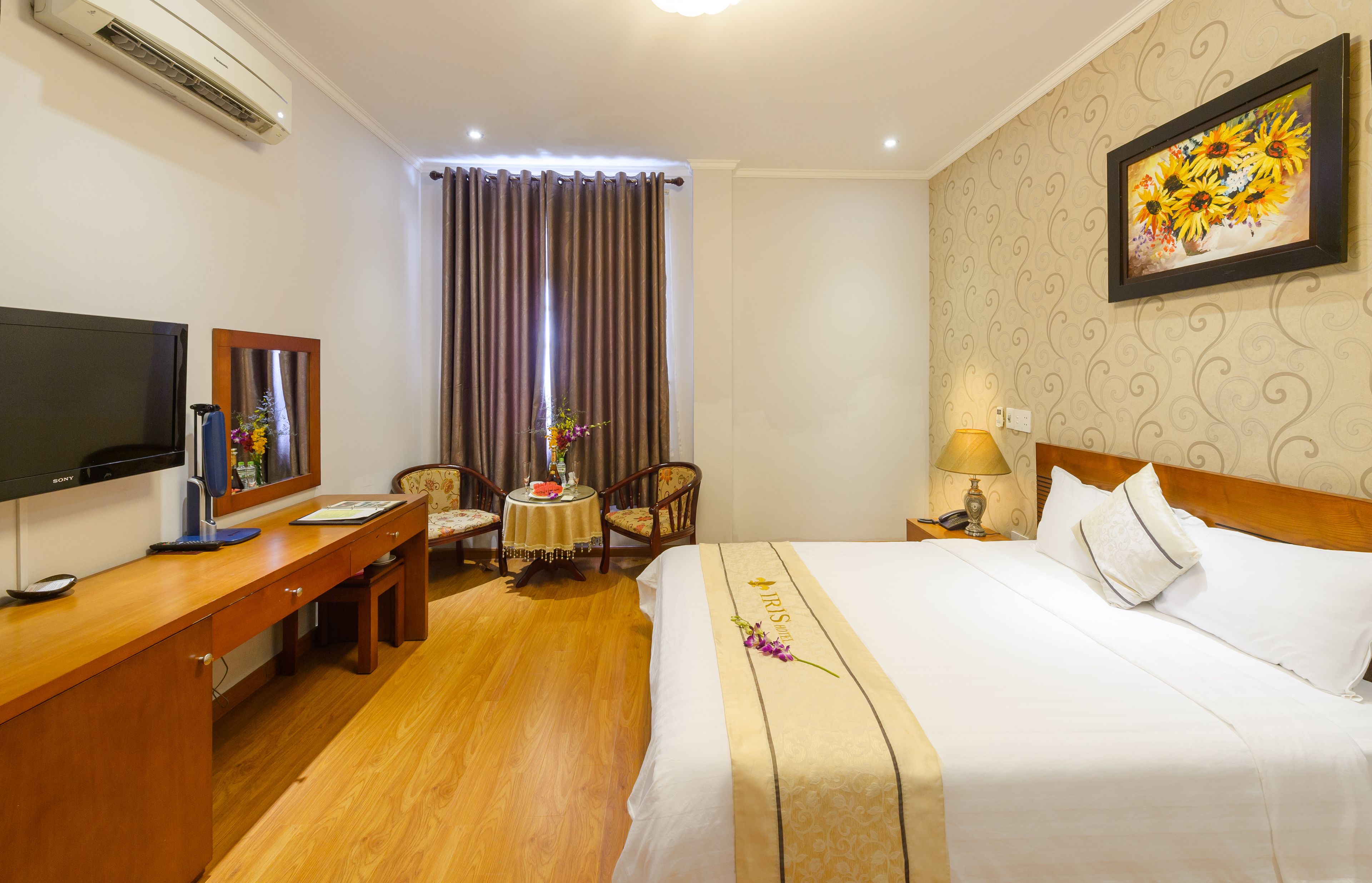 A hotel room with a bed, a desk with a computer, a chair, a table with a vase, and a wall-mounted TV.