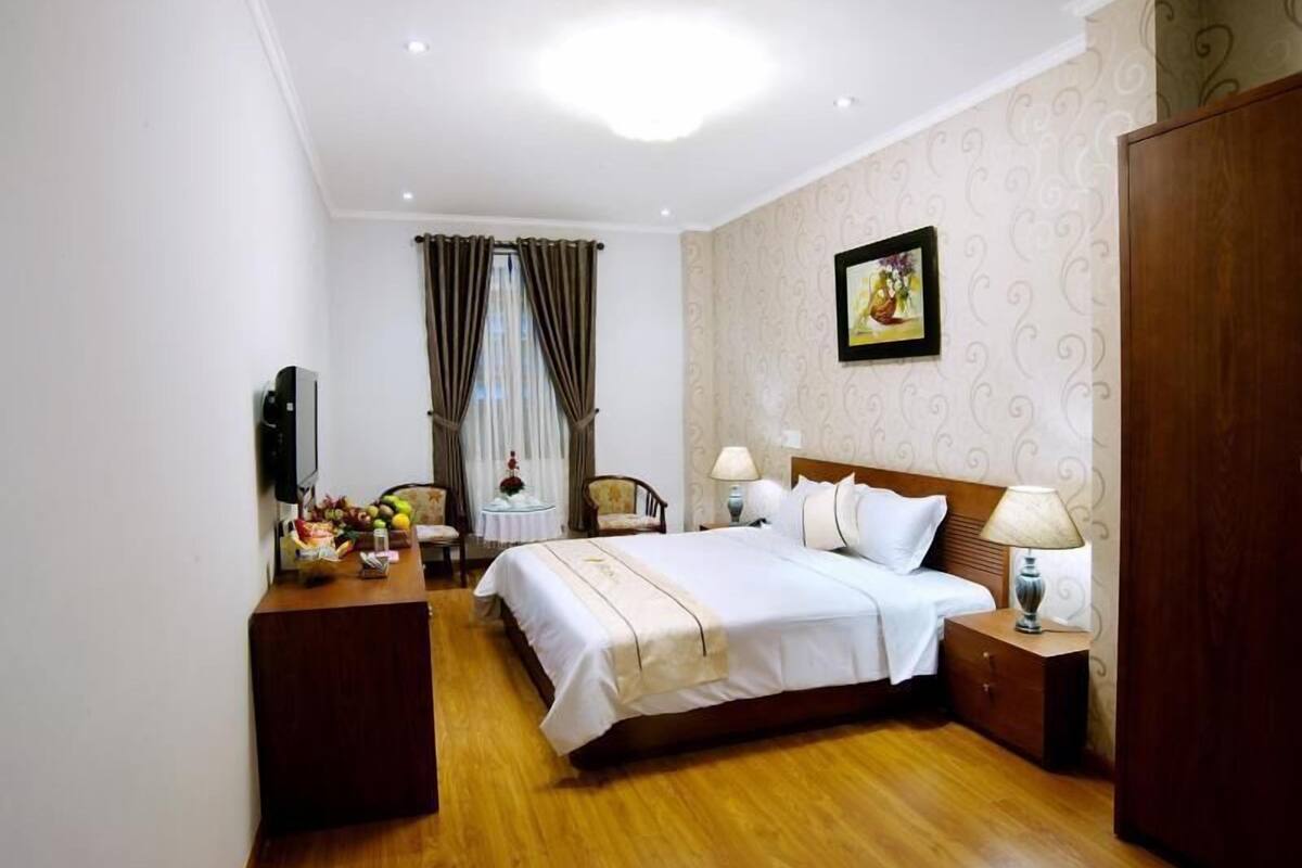 Una habitación de hotel con cama, mesitas de noche, un escritorio con silla, televisión y ventana con cortinas.