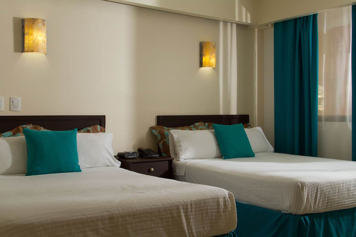 Habitación de hotel con dos camas, cada una con ropa blanca y almohadas turquesa, una mesita de noche con teléfono y apliques de pared.