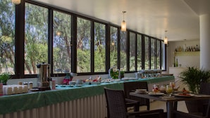 A breakfast bar with a view of trees through large windows, a coffee maker, and a variety of breakfast items.