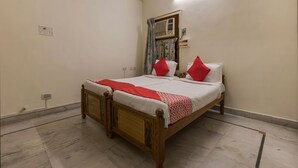 Een slaapkamer met een houten bed, wit beddengoed, rode kussens, een rode en witte geborduurde plaid, een raam met gordijnen en een luchtverkoeler aan de muur.
