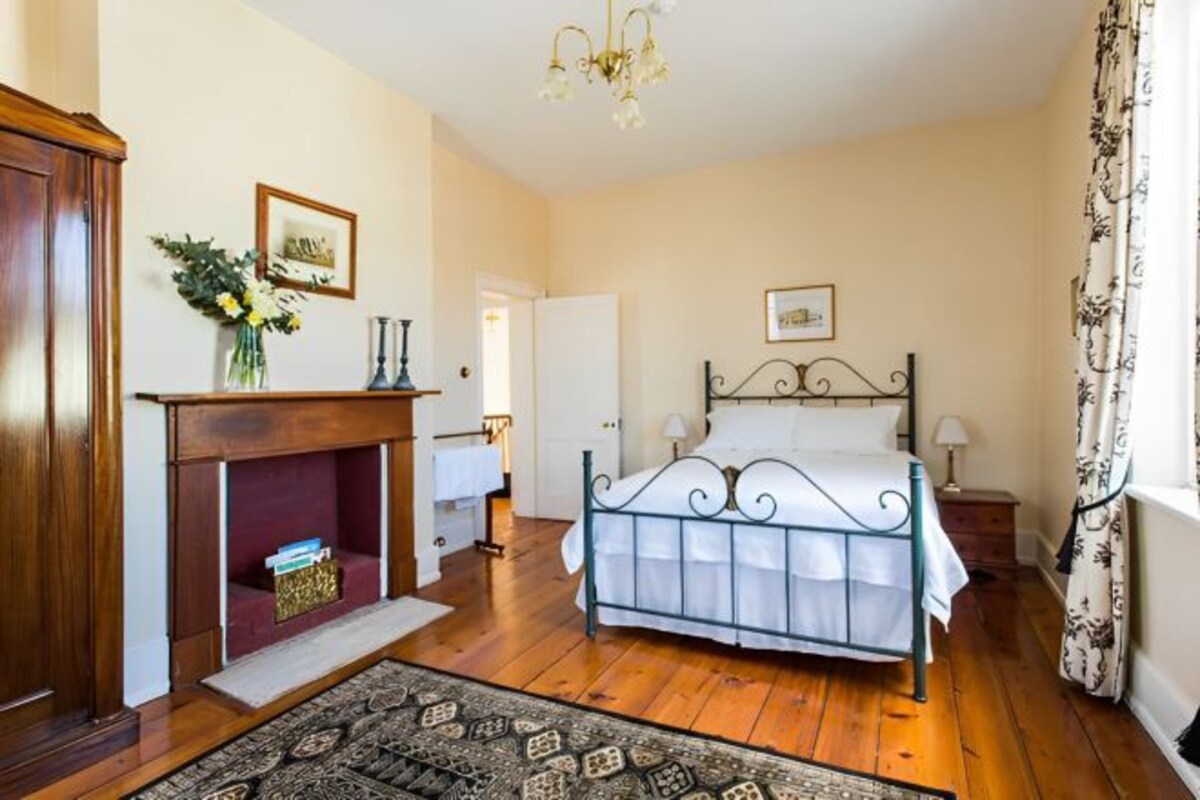 A bedroom with a wooden floor, a metal bed frame, a fireplace, and a chandelier.