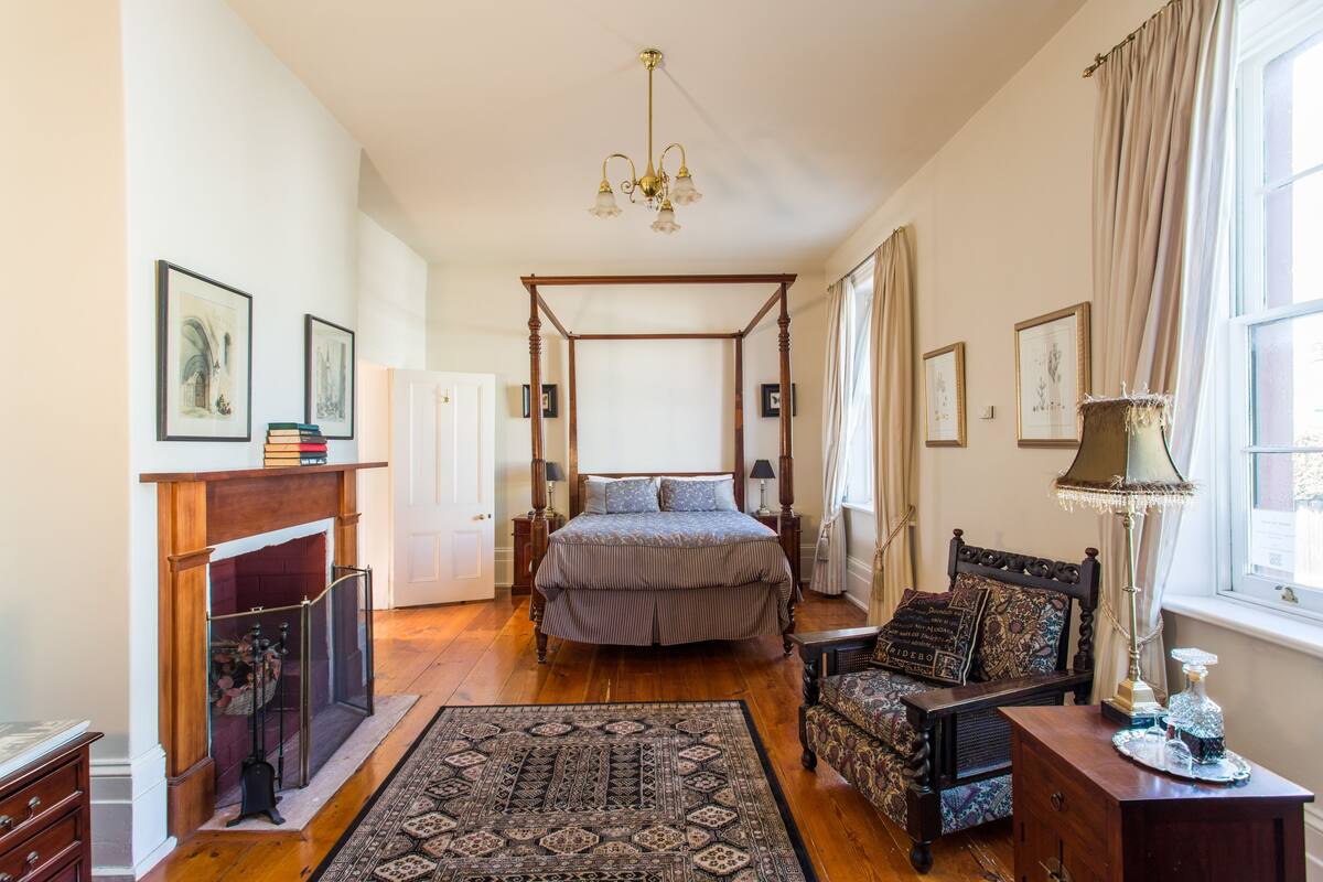 Une chambre à coucher de style traditionnel, avec un lit à baldaquin, une cheminée, un lustre et une grande fenêtre agrémentée de rideaux.