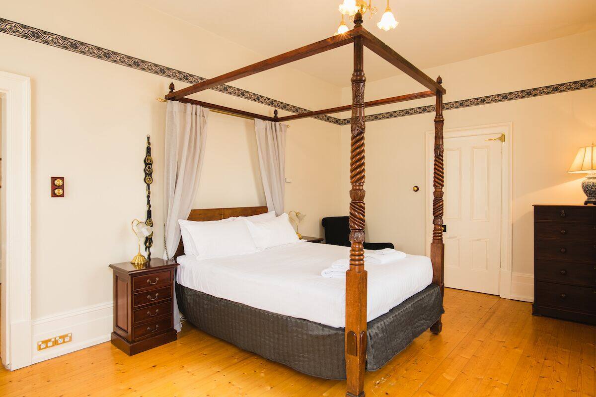 Un lit à baldaquin avec une structure en bois et un ciel de lit, une table de chevet avec une lampe, et une commode sur la droite.