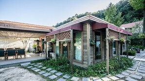 A wooden cabin with a covered patio area, stone pathway, and surrounding greenery.