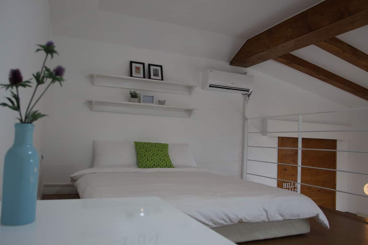 Een slaapkamer met een bed, witte muren, een houten balk in het plafond, een witte nachtkastje, een blauwe vaas en een kussen met een groen patroon.