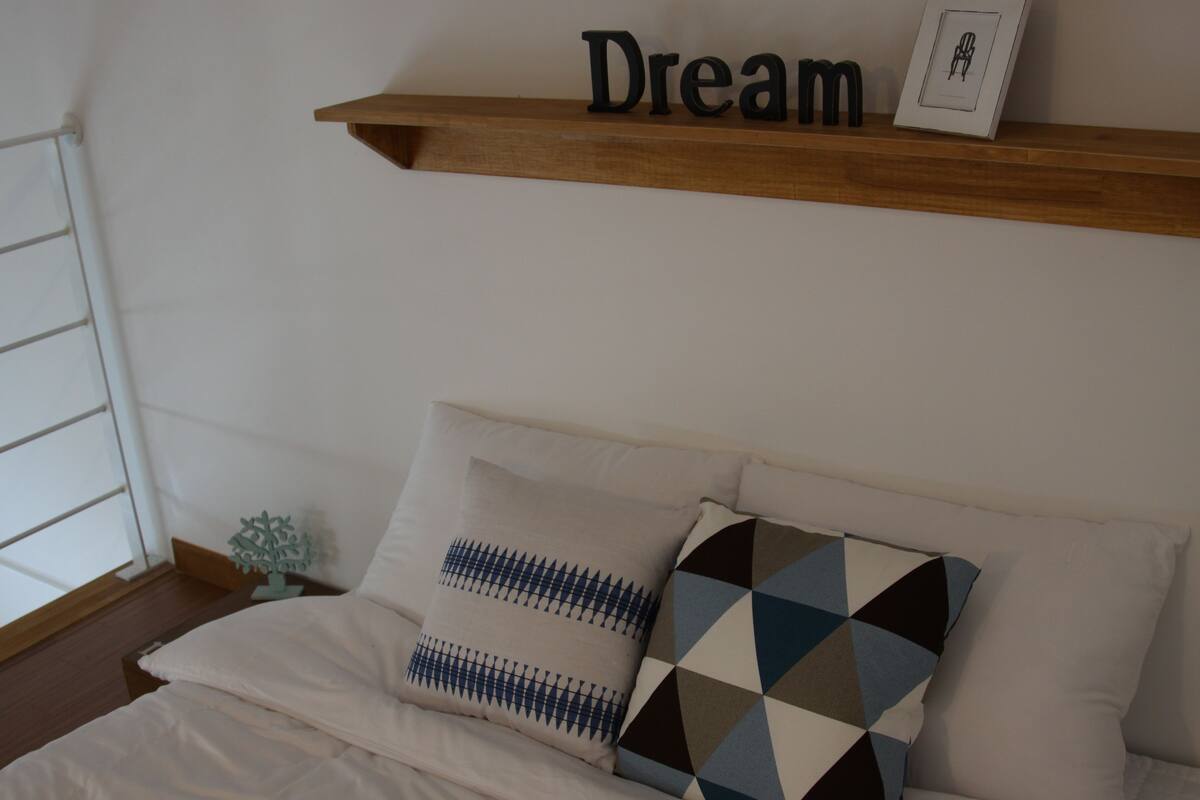 Een netjes opgemaakt bed met twee kussens, een houten plank erboven met een ingelijste foto en de tekst 'Dream', en een kleine decoratieve sneeuwvlok op een nachtkastje.