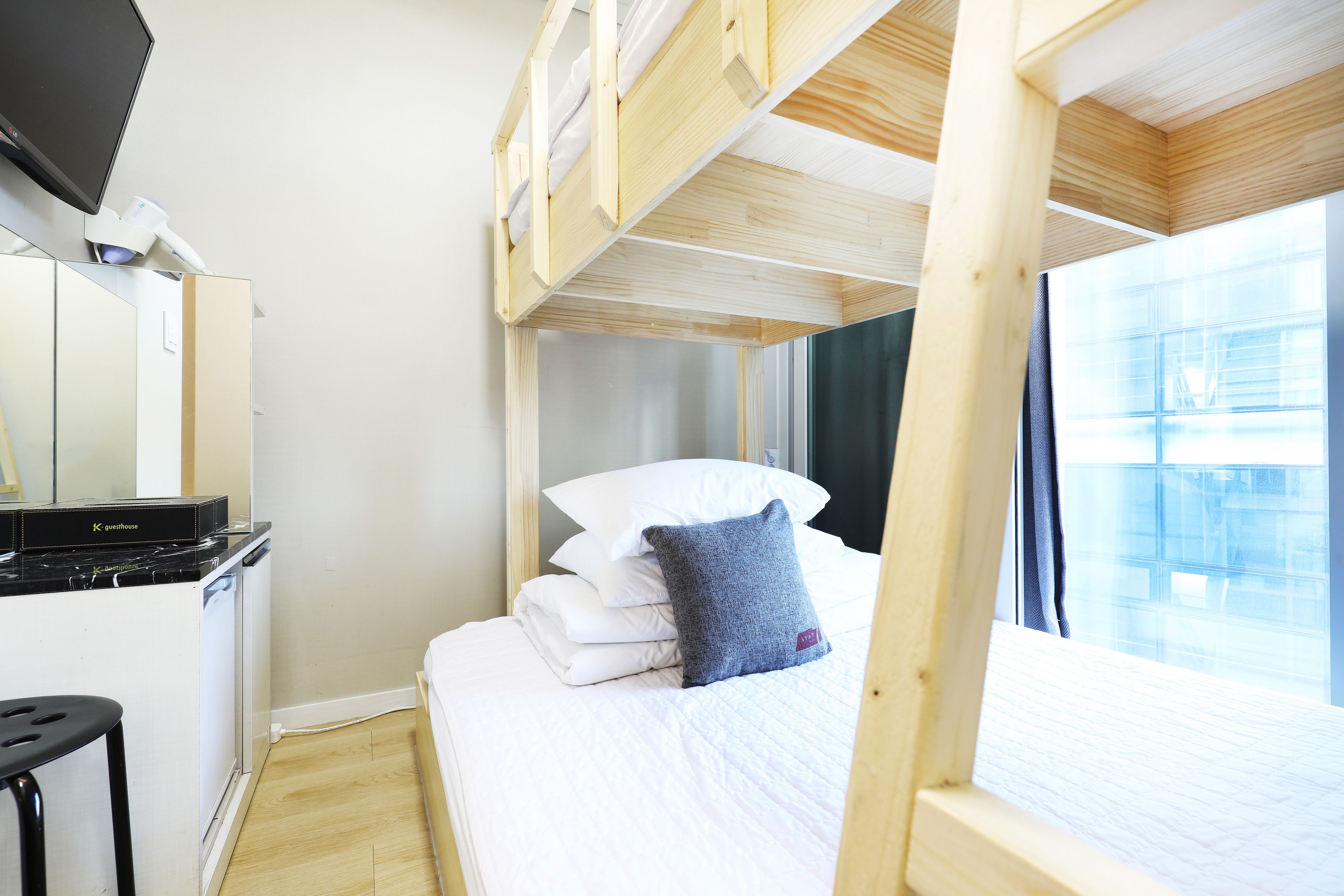 A bunk bed with white bedding and a blue pillow, a small desk with a computer, and a window with curtains.