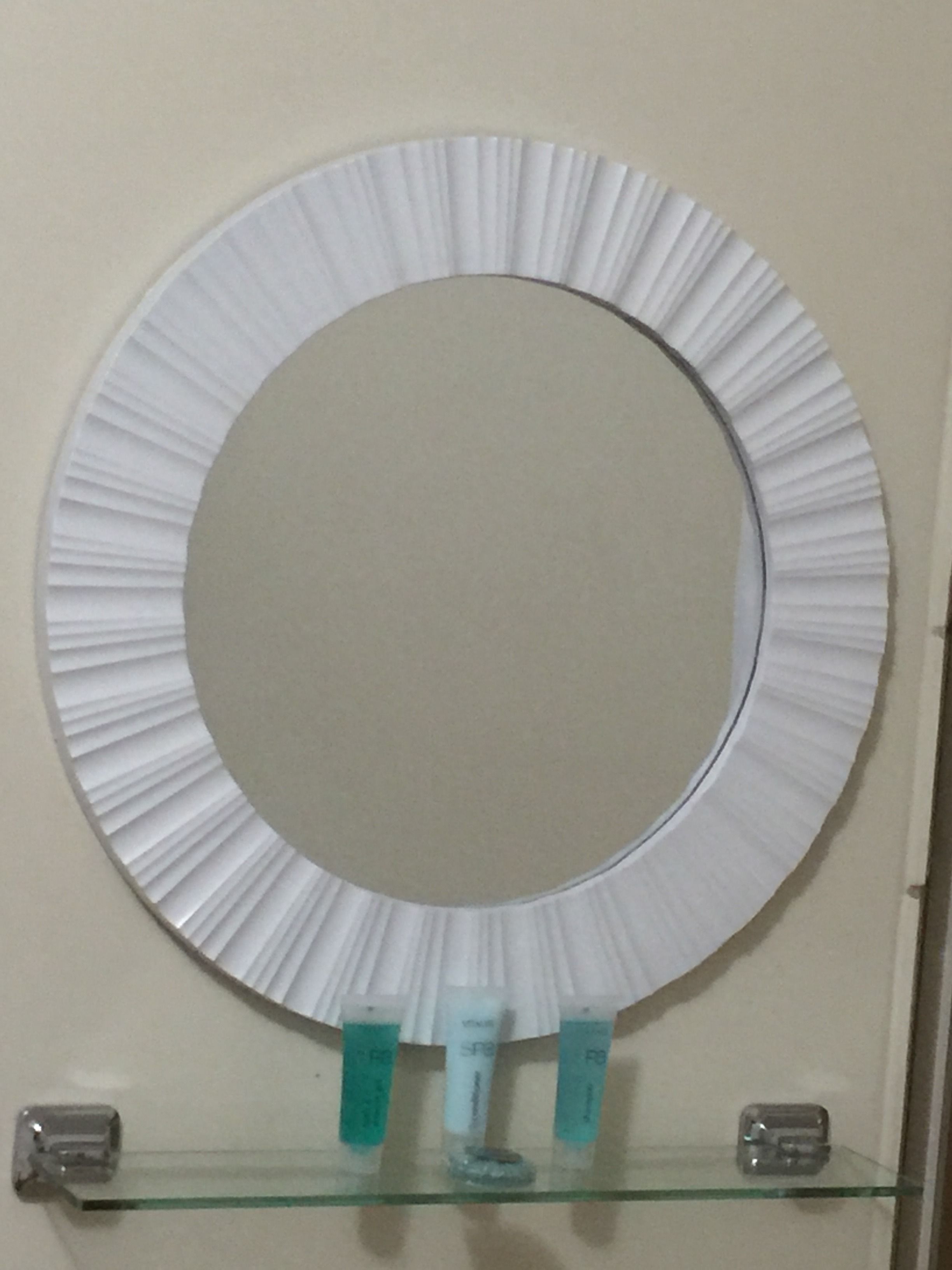 A round mirror with a pleated design mounted on a wall above a glass shelf with three small containers.