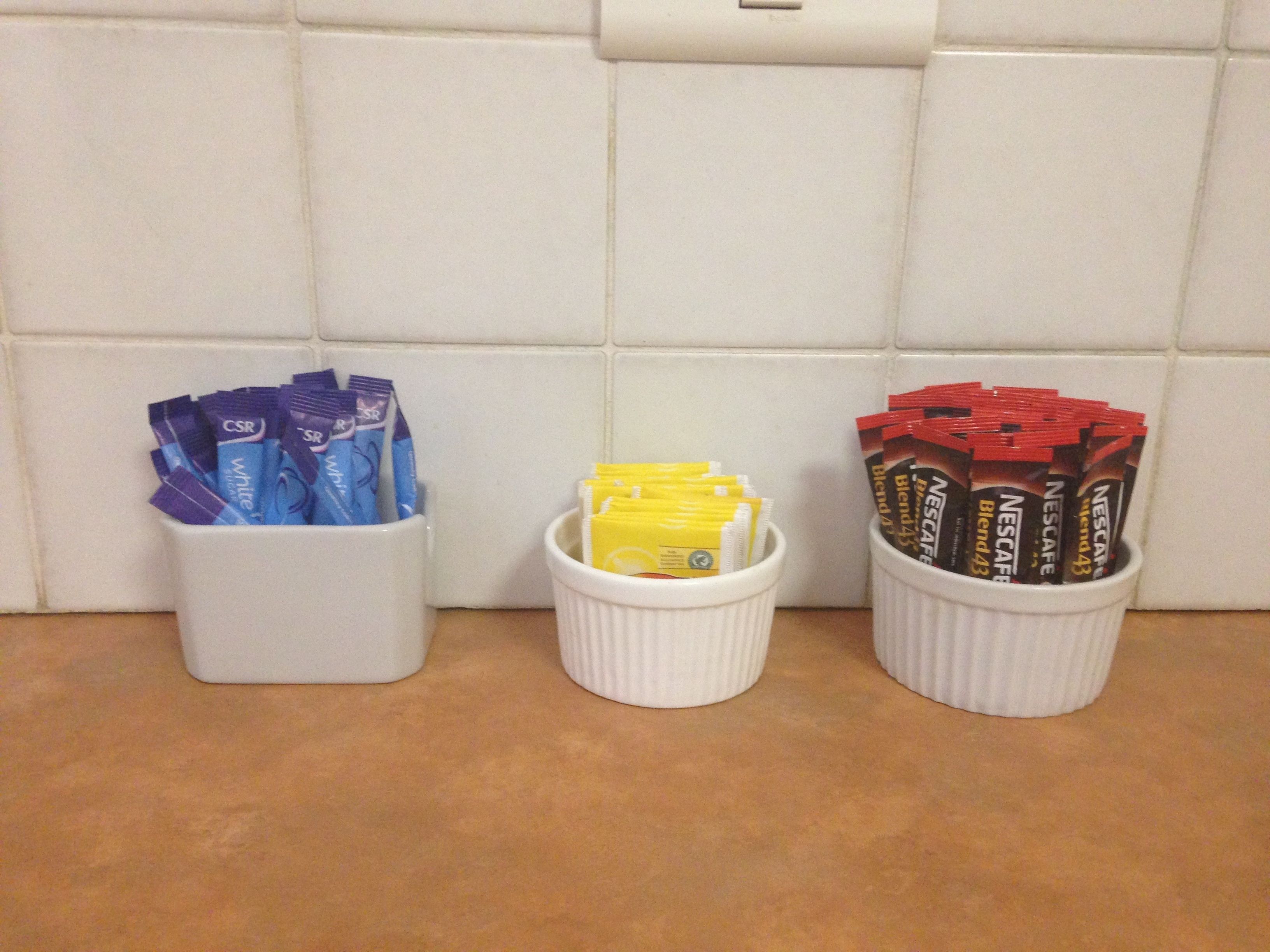 Três recipientes brancos em uma bancada de cozinha, cada um contendo cápsulas de café de cores diferentes.