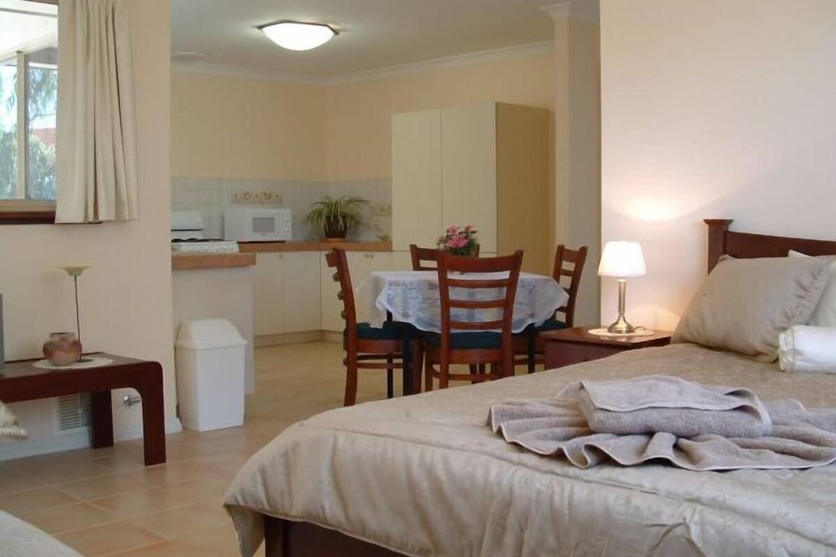 A hotel room with a bed, bedside table, lamp, and a view of the kitchen and dining area.