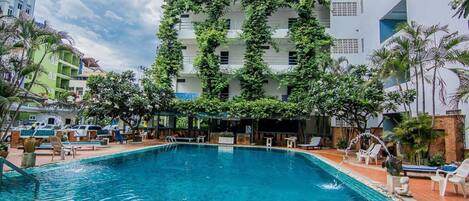 Piscina com espreguiçadeiras e guarda-sóis, rodeada por edifícios com áreas verdes nas fachadas.