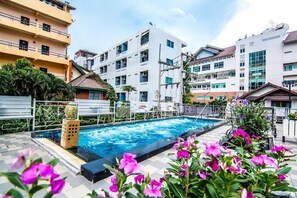 Ein Swimmingpool, umgeben von einem Garten mit rosa Blumen, neben einem mehrstöckigen Wohngebäude mit Balkonen.