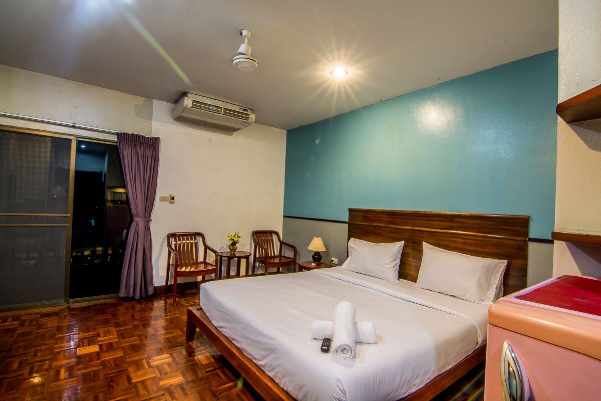 A hotel room with a wooden floor, a bed with white linens, a wooden headboard, a nightstand with a lamp, a chair, and a small table with flowers.
